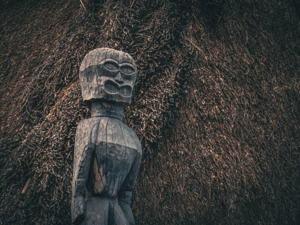 Puʻuhonua o Hōnaunau National Historical Park, Hōnaunau, Hawai'i (photo August 21, 2021): recreated ki'i (divinity statue) against Hale o Keawe (thatched mortuary temple built by or for ruler Keawe-i-kekahi-aliʻi-o-ka-moku c.1700, last restored 2004).